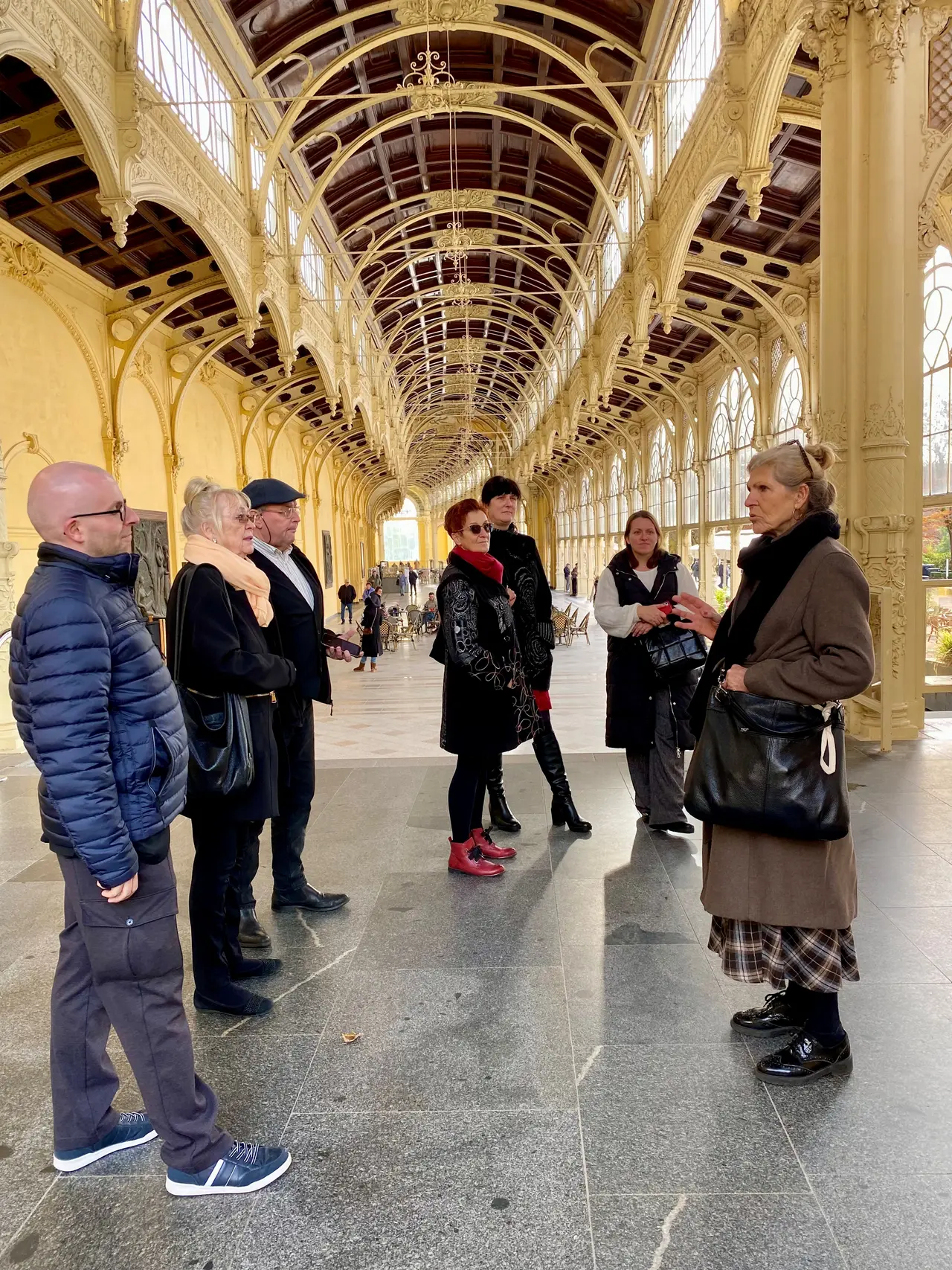 Eine Gruppe von Menschen steht mit einer Reiseführerin in einer Kolonade in Marienbad und lauscht ihren Erklärungen