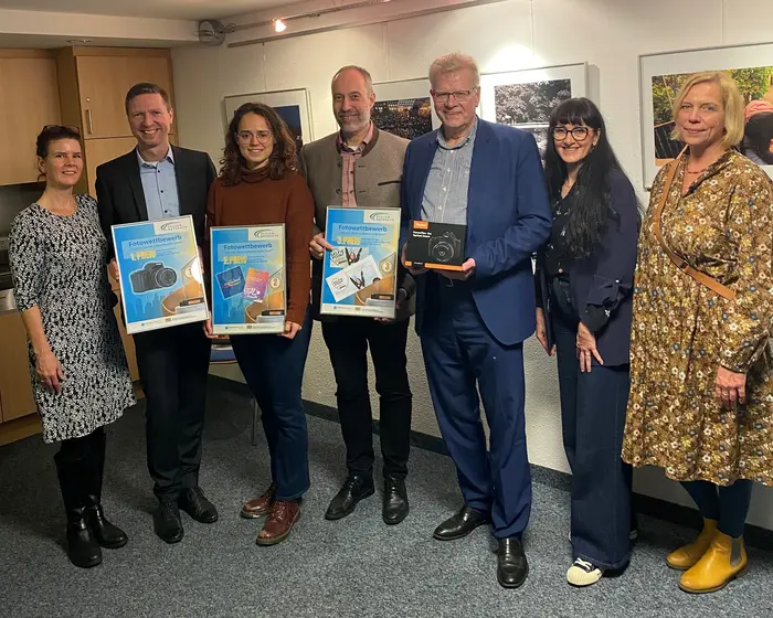 7 Personen stehen vor Fotografien und halten Preise für einen Wettbwerb hoch.